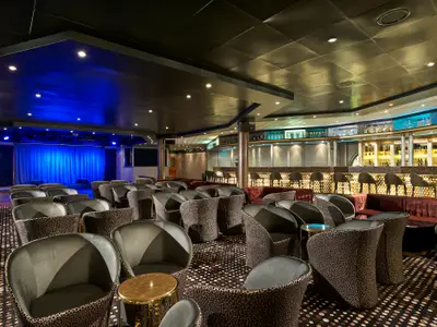 Innenansicht der Bar-Lounge "Sun Bliss Lounge" an Bord der Norwegian Sun. Die Lounge hat eine Bühne mit blauer Beleuchtung und eine lange Bar mit Stühlen. Elegante Sessel prägen den Raum.