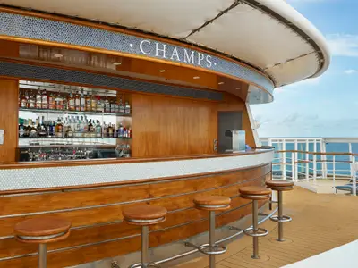 Die Champs Bar an Bord der Norwegian Sun zeigt eine Holztheke mit Barhockern und Spirituosenregalen. Im Hintergrund ist das offene Meer sichtbar.