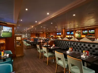 Innenansicht der Sportsbar an Bord der Norwegian Sun. Der Raum bietet Holztische, Stühle, dunkle Lederbänke, eine Bar und mehrere Bildschirme, die Golf zeigen.