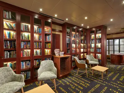 Eine gemütliche Bibliothek an Bord der Norwegian Sun mit hohen, beleuchteten Holzregalen voller Bücher, bequemen Sesseln, kleinen Tischen und einem gemusterten Teppich. Im Hintergrund sind weitere Regale und ein abgerundeter Tresen zu sehen.