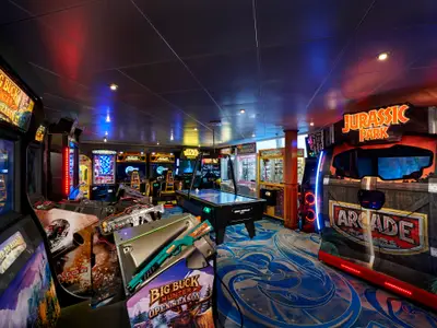 Ein lebhaftes Spielzimmer der Norwegian Sun mit bunten Lichtern und zahlreichen Spielautomaten wie 'Big Buck Hunter', 'Jurassic Park' und einem Airhockey-Tisch.