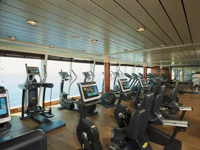 Ein geräumiges Fitnesscenter an Bord der Norwegian Sun, ausgestattet mit modernen Cardio- und Kraftgeräten. Große Fensterfronten bieten einen weiten Blick auf das offene Meer, während die Decke mit integrierten Leuchten für helle Beleuchtung sorgt. Der Raum wirkt gepflegt und einladend.