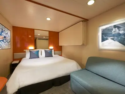 Kabine auf der Norwegian Sun mit Doppelbett, blauen Kissen und einem Sofa. Zwei gerahmte Kunstwerke haengen an den Waenden. Ein weiteres Bett ist hochgeklappt.