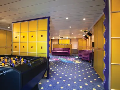 Innenansicht des Jugendbereichs an Bord der Norwegian Sun. Ein Foosballtisch steht vorne, dahinter gelbe Wände und eine violette Couch. Der Boden hat einen blauen Teppich mit weissen Punkten und bunten Flecken.