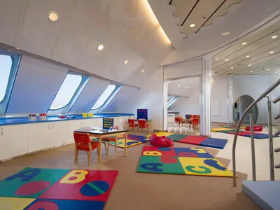 Ein heller Kinderbereich an Bord der Norwegian Sun mit schrägen Fenstern, bunten Spielmatten, Tischen mit Stühlen und Spielzeug wie Kegeln und Connect Four. Eine Wendeltreppe ist rechts zu sehen.