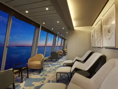 Eine moderne Lounge an Bord der Norwegian Sun mit bequemen Liegestühlen und Sesseln. Große Fensterfronten bieten einen weiten Blick auf das Meer und den Horizont während der Dämmerung mit blauem Himmel und rosa Lichtern. Gerahmte Kunstwerke schmücken die Wände.