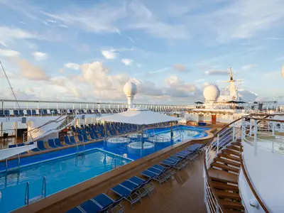 Das sonnige Pooldeck der Norwegian Sun zeigt blaue Pools, Whirlpools und Reihen von Liegestühlen. Im Hintergrund sind das Meer und ein leicht bewölkter Himmel zu sehen.