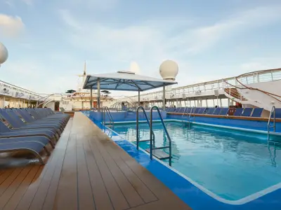 Sonniges Pooldeck der Norwegian Sun mit zwei blauen Pools, Whirlpools und vielen Liegestühlen. Decks sind menschenleer, der Himmel ist klar.
