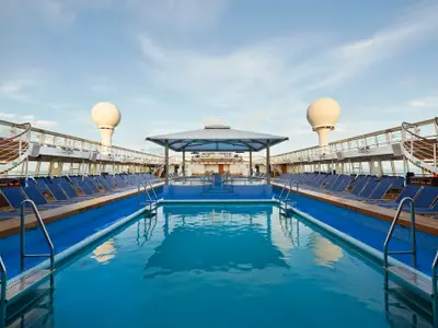 Das Hauptpooldeck an Bord der Norwegian Sun bei blauem Himmel. Zwei Pools sind von blauen Liegestühlen gesäumt. Eine Überdachung und zwei große, weiße Radome sind sichtbar.