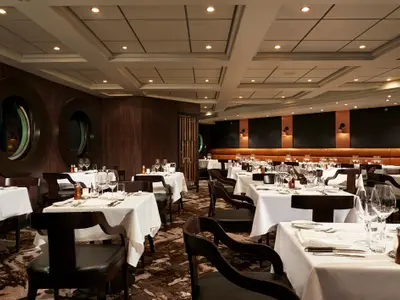 Elegantes Spezialitätenrestaurant an Bord der Norwegian Sun. Weiss gedeckte Tische mit Gläsern und Besteck. Das Interieur zeigt dunkle Holzvertäfelungen, runde Fenster und eine lange braune Lederbank.