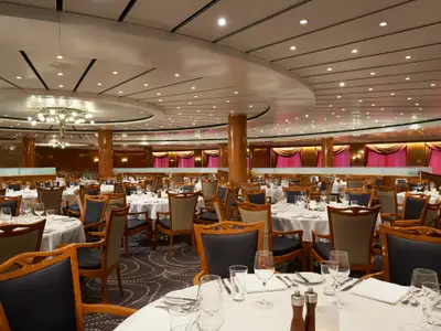 Ein geräumiger Speisesaal, das Four Seasons Restaurant, an Bord der Norwegian Sun, mit zahlreichen, elegant weiss gedeckten Tischen, Holzstühlen und roten Vorhängen. Eine grosse Kronleuchter hängt von der Decke.