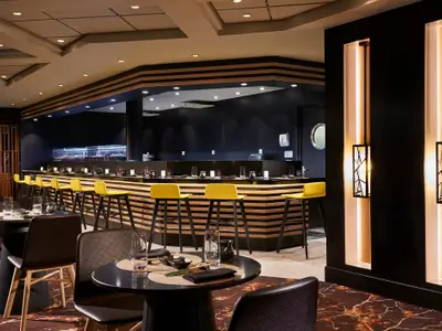 Sushi-Restaurant Ginza an Bord der Norwegian Sun. Lange Bar mit gelben Hockern und dunklem Dekor. Tische sind gedeckt.