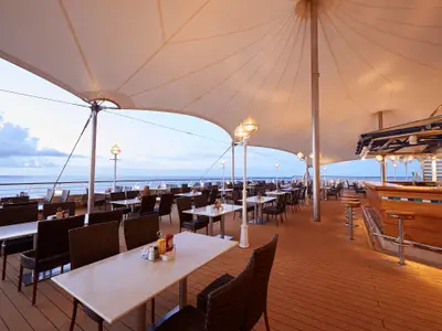 Aussenrestaurant 'The Great Outdoors' an Bord der Norwegian Sun. Tische, Stühle und Holzbar unter Sonnensegel. Meer und Himmel bei Abendstimmung, Lampen erhellen die Szene.