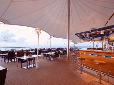 Weitwinkelaufnahme des überdachten Außenrestaurants und einer voll ausgestatteten Bar an Bord der Norwegian Sun, mit zahlreichen leeren Tischen und Stühlen und Blick auf das ruhige Meer im Abendlicht.