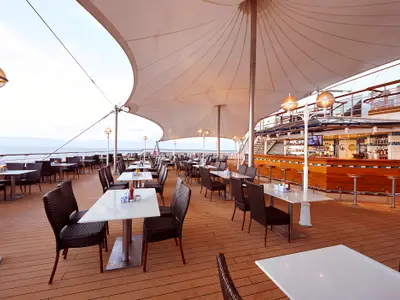 Grosszügiges Aussendeck-Restaurant und Bar an Bord der Norwegian Sun. Viele Tische und Stühle unter einem Sonnensegel, eine lange Bar mit Hockern, dahinter das weite Meer.