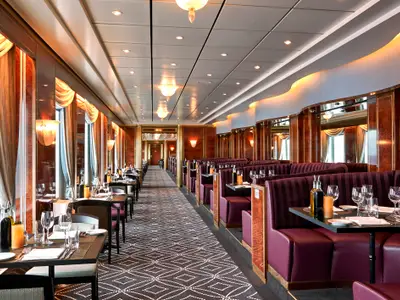 Innenansicht des Spezialitätenrestaurants an Bord der Norwegian Sun. Gedeckte Tische mit Gläsern und Besteck. Links Fenster, rechts lila Bankett-Sitze. Klassisches Design mit Holz und warmem Licht.