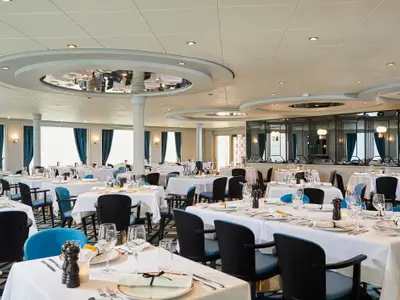Ein elegant gedecktes Restaurant an Bord der Norwegian Sun mit zahlreichen Tischen und blauen Stühlen. Der Raum ist hell und modern gestaltet mit großen Fenstern.