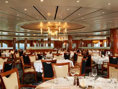 Eleganter Speisesaal der Norwegian Sun. Viele gedeckte Tische, stilvolle Stühle, Holzsäulen und Kronleuchter. Fenster im Hintergrund zeigen das Meer.