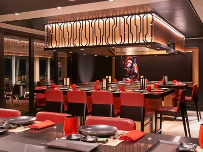Spezialitätenrestaurant Teppanyaki an Bord der Norwegian Sun. Mehrere Tische mit rotem Lederstühlen um Grillplatten. Japanische Kunst an der Wand, darunter eine Geisha-Darstellung. Moderne Beleuchtung und Einrichtung.