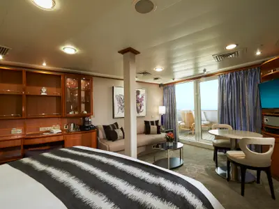 Blick in eine helle, elegante Suite an Bord der Norwegian Sun mit einem grossen Bett, einem Sofa, einem Esstisch und einem Balkon mit Stühlen und Meerblick. Holzmöbel und moderne Kunst schmücken den Raum.
