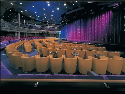 Innenansicht der Stardust Lounge an Bord der Norwegian Sun. Goldbraune Sessel und kleine Tische vor einer blau-violett beleuchteten Bühne mit funkelndem Vorhang. Oben weitere Sitzreihen.