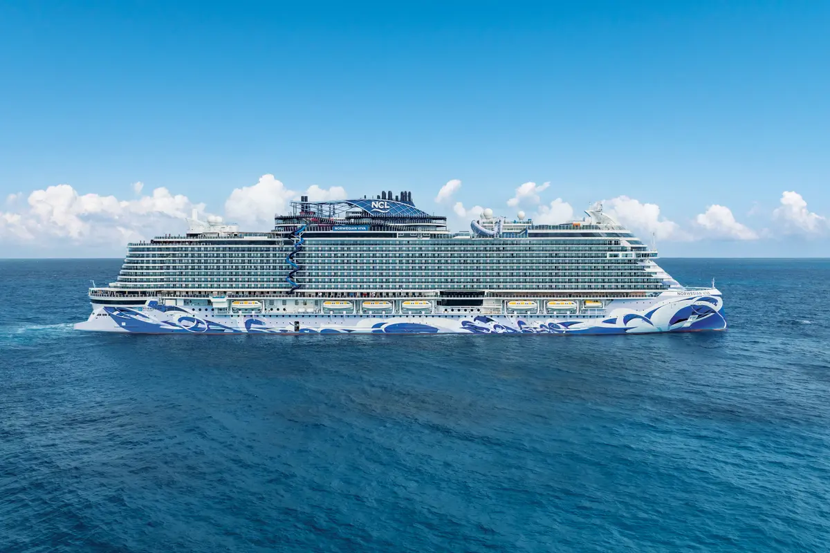 Aussenansicht der Norwegian Viva auf dem Meer. Das grosse Kreuzfahrtschiff mit einzigartiger blau-weisser Rumpfbemalung gleitet unter blauem Himmel mit Wolken durchs Wasser. Keine Personen sichtbar.