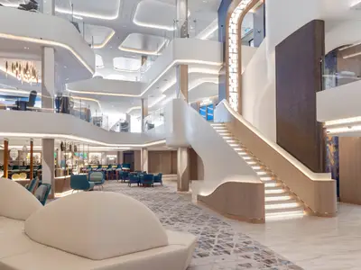 Das helle Atrium der Norwegian Viva mit einer eleganten, geschwungenen Holztreppe und integrierter Beleuchtung. Mehrere Lounges, eine Bar und moderne Kunstinstallationen sind zu sehen.