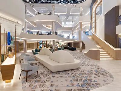 Ein elegantes, modernes Atrium an Bord der Norwegian Viva mit einer grossen geschwungenen Treppe, bequemen Lounge-Sesseln und einem Teppich mit blau-braunem Muster. Mehrere Etagen sind sichtbar.