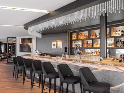 Eine moderne und elegante Bar-Lounge an Bord der Norwegian Viva. Im Vordergrund eine lange, beleuchtete Theke mit dunklen Barhockern. Eine beeindruckende Kristall-Lichtinstallation hängt über der Theke. Im Hintergrund sind Regale mit Spirituosen und Kunstwerke zu sehen.