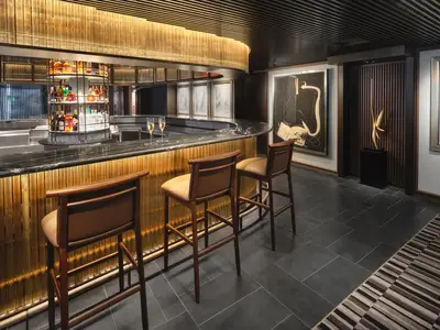 Eine luxuriöse Bar an Bord der Norwegian Viva mit schwarzem Marmortresen, goldener Rippenverkleidung und bequemen Barhockern. Zwei Champagnergläser stehen auf dem Tresen. Im Hintergrund sind moderne Kunstwerke zu sehen.