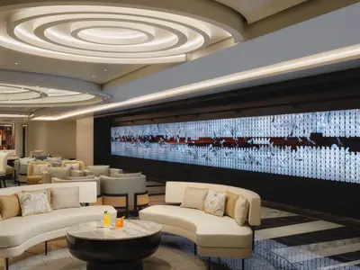 Eine stilvolle Bar-Lounge an Bord der Norwegian Viva. Im Vordergrund geschwungene cremefarbene Sofas mit Zierkissen und ein runder Couchtisch mit Cocktails. Eine lange, beleuchtete LED-Wand mit abstrakten Mustern dominiert die rechte Wand. Elegante Deckenbeleuchtung erhellt den Raum.