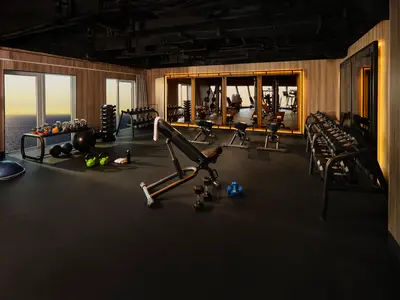 Fitnessstudio an Bord der Norwegian Viva. Meerblick durch Fenster. Ausgestattet mit Hanteln, Kettlebells, Trainingsbank. Holzvertäfelte Wände und beleuchtete Spiegel.
