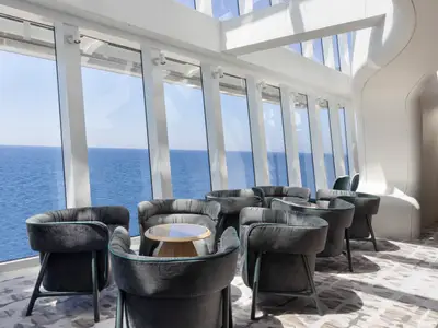 Lounge an Bord der Norwegian Viva. Große Fensterfront mit Meerblick. Dunkelgraue Sessel und kleine Tische in hellem, modernem Ambiente. Viel Tageslicht.
