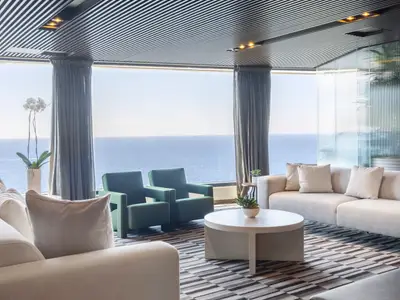 Moderne Lounge an Bord der Norwegian Viva mit bodentiefen Fenstern und Meerblick. Helle Sofas, dunkelgrüne Sessel und ein runder Couchtisch prägen den Raum.
