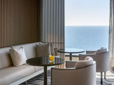 Moderne Lounge an Bord der Norwegian Viva. Beiges Sofa, Tische mit Cocktail, Sessel und vertikale Lamellen an der Wand. Weitreichender Meerblick durch das Fenster.