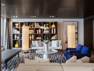 Die stilvolle Haven Lounge an Bord der Norwegian Viva. Ein großer, dunkler Bücherschrank voller Vasen und Bücher dominiert den Raum. Weiße Sofas, blaue Sessel, ein heller Marmortisch und ein gestreifter Teppich bilden eine moderne Sitzgruppe. Zwei hohe Stehlampen beleuchten den Bereich.
