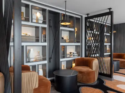 Eine moderne Lounge an Bord der Norwegian Viva. Cognacfarbene Samtsessel stehen vor einer Wand mit dekorativen Regalen voller Kunstobjekte. Eine schwarze Pendelleuchte ist sichtbar.