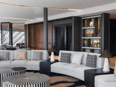 Eine luxuriöse, helle Lounge an Bord der Norwegian Viva mit großen Fenstern und Blick auf eine Landschaft. Helle Sofas, kunstvolle Tische und dekorierte Regale prägen den Raum.