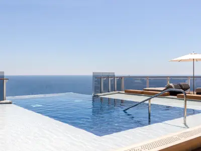 Infinity-Pool der Norwegian Viva auf dem Sonnendeck. Das blaue Wasser geht optisch ins Meer über. Sonnenliegen, ein Sonnenschirm und Glasbrüstungen sind sichtbar. Blauer Himmel.