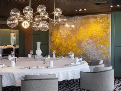 Elegantes Restaurant an Bord der Norwegian Viva. Ein runder Tisch, weiß gedeckt, mit Gläsern und modernen Dekorationen. Ein goldenes Wandkunstwerk und ein Kronleuchter schmücken den Raum.