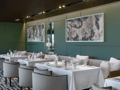 Innenansicht eines eleganten Restaurants an Bord der Norwegian Viva. Mehrere weiss gedeckte Tische sind bereit, umgeben von gepolsterten Bänken und Stühlen. An der grünen Wand hängen moderne Kunstwerke.