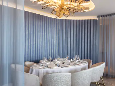 Ein elegantes Restaurant an Bord der Norwegian Viva. Ein ovaler Tisch ist festlich gedeckt mit Geschirr, Besteck und Gläsern. Eine geschwungene Wand mit blauen Vorhängen bildet den Hintergrund. Goldene Deckenleuchte.
