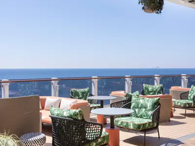 Sonniger Loungebereich an Deck der Norwegian Viva mit Meerblick. Korbsessel und Sofas mit tropisch gemusterten Kissen laden zum Entspannen ein. Ein weiteres Schiff ist am Horizont sichtbar.