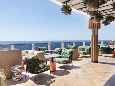 Eine weitläufige Outdoor-Lounge an Bord der Norwegian Viva mit gemütlichen Sitzgruppen in Grüntönen und orangen Akzenten. Der Bereich ist teilweise überdacht und bietet einen weiten Blick auf das offene Meer unter blauem Himmel. Hängepflanzen schmücken die Decke.