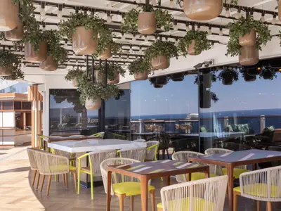 Ein helles Restaurant an Bord der Norwegian Viva mit stilvollen Tischen und Stühlen. Zahlreiche Pflanzen hängen von der Decke, während große Glasfenster einen weiten Blick auf das blaue Meer bieten.