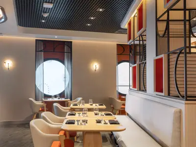 Innenansicht eines eleganten Restaurants an Bord der Norwegian Viva. Mehrere Holztische sind mit Gläsern und dunklen Platzsets gedeckt. Helle Polstersessel und eine lange Sitzbank säumen den Raum. Zwei große runde Bullaugenfenster mit hellen Vorhängen lassen Tageslicht herein. Die Wände sind in warmen Cremetönen gehalten, und dekorative Wandleuchten spenden zusätzliches Licht. Eine Trennwand mit roten Akzenten und schwarzen Gitterelementen im japanischen Stil ist rechts sichtbar.