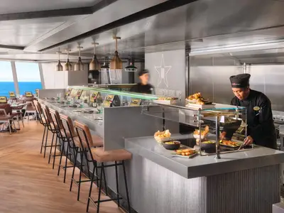 Blick in das Spezialitätenrestaurant "Food Republic" an Bord der Norwegian Viva, wo ein Koch Speisen an einem modernen Tresen zubereitet. Im Hintergrund ist Meerblick durch große Fenster zu sehen.