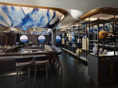 Ein elegantes Teppanyaki-Restaurant namens Hasuki an Bord der Norwegian Viva, mit dunklen Holztheken und Stühlen. Blaue Kunstwerke schmücken die Decke, während runde Bullaugen einen Blick auf das Meer bieten. Eine Regalwand mit farbigen Keramikvasen dient als Raumteiler.