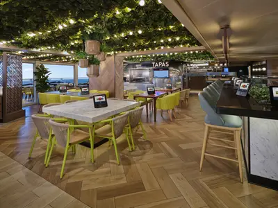 Modernes Restaurant an Bord der Norwegian Viva. Deckenbegrünung, Lichter, Meerblick. Sitzbereiche mit gelben Stühlen. Bar mit Hockern und Bestellterminals.