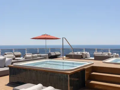 Sonnendeck der Norwegian Viva. Zwei Whirlpools, Lounge-Möbel und roter Sonnenschirm. Blick auf weites blaues Meer und klaren Himmel, durch Glasbrüstung getrennt.